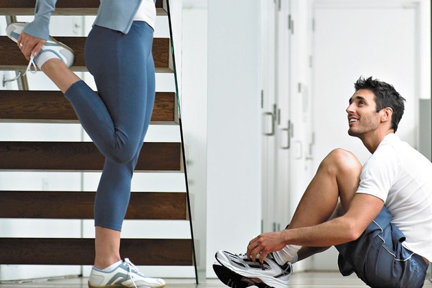 A woman stretching before her run, and a man sitting and tying his shoes looking up at her